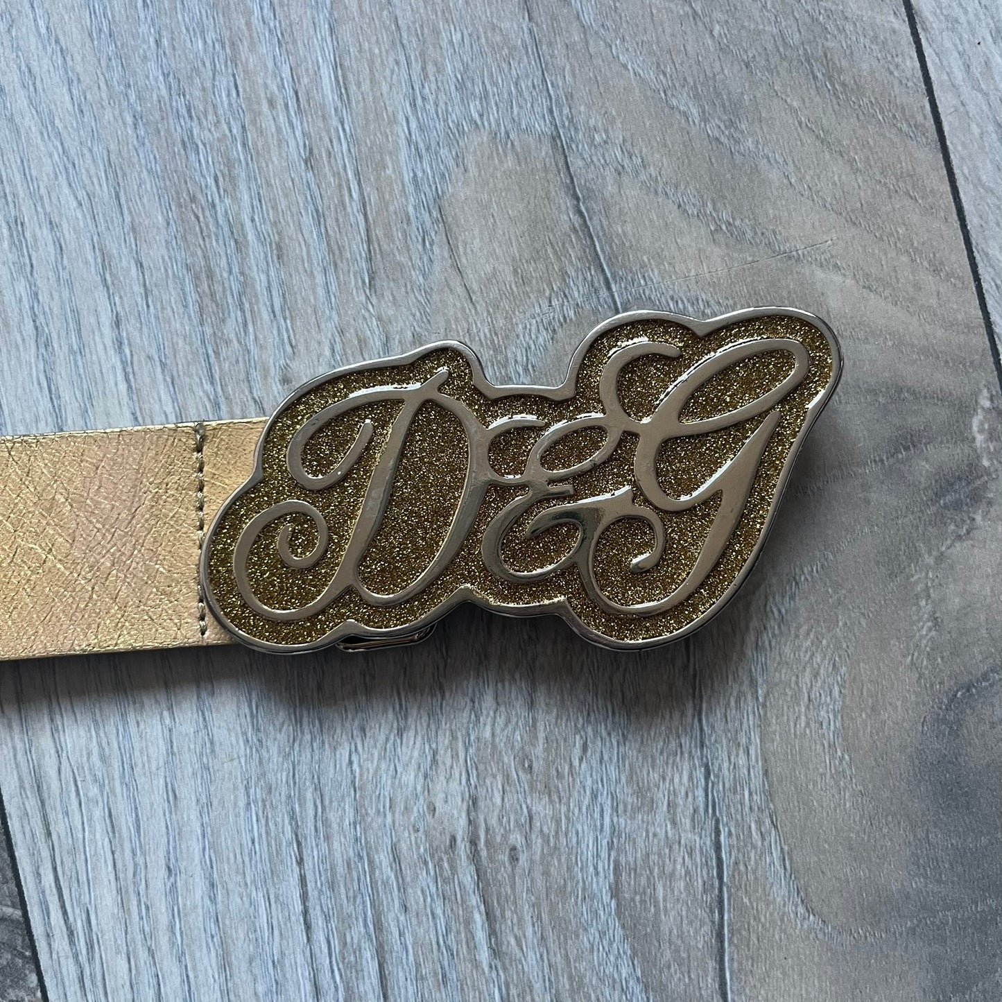 The Dolce & Gabbana 2000’s Gold Tone D&G Logo Belt is made from metallic gold leather and features a large D&G logo buckle shaped directly as the brand’s initials. The buckle uses a cursive D&G logo set over a glittery gold background, framed by a solid gold-tone metal outline. The entire buckle and belt maintain a consistent gold color scheme typical of Dolce & Gabbana accessories from the 2000s.