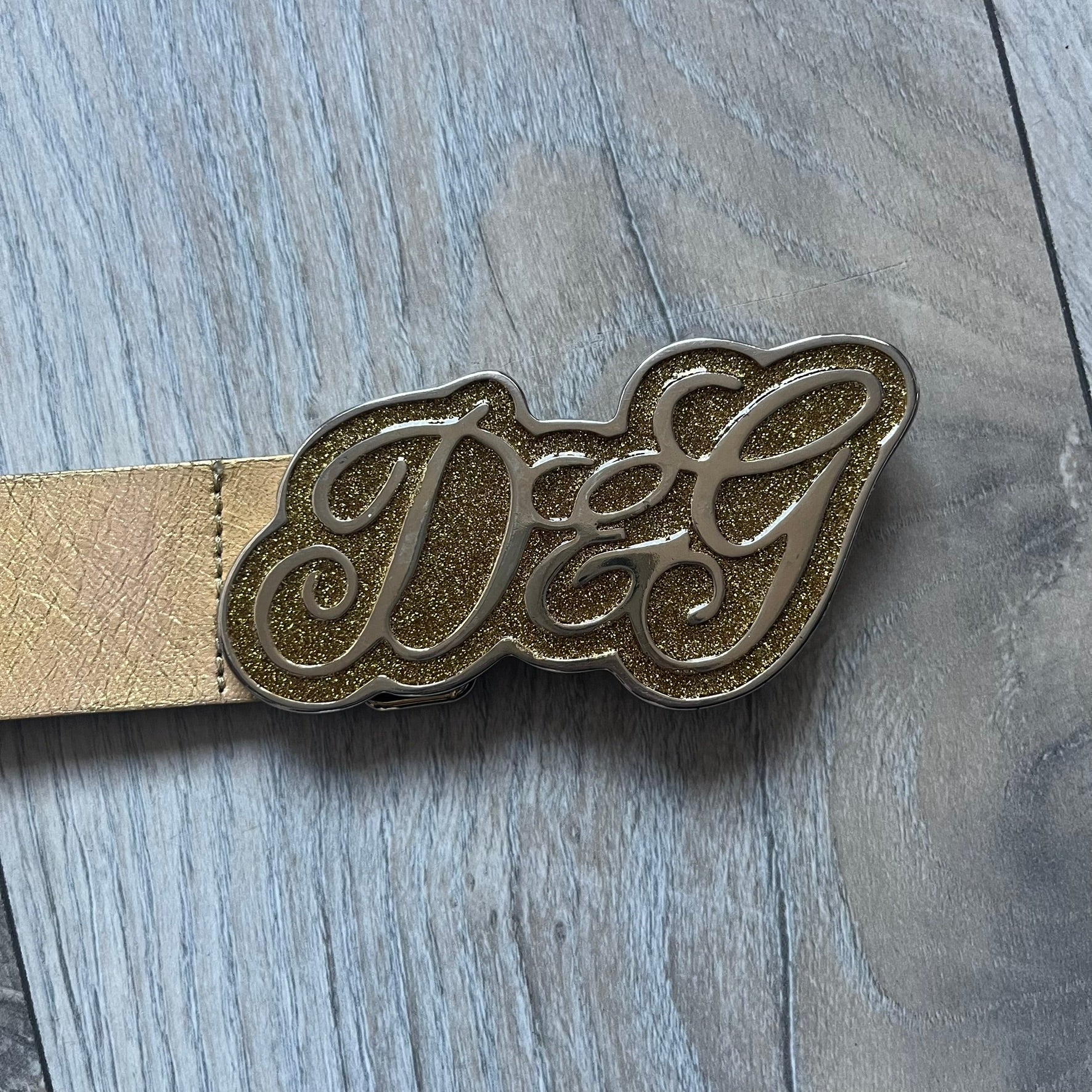 The Dolce & Gabbana 2000’s Gold Tone D&G Logo Belt is made from metallic gold leather and features a large D&G logo buckle shaped directly as the brand’s initials. The buckle uses a cursive D&G logo set over a glittery gold background, framed by a solid gold-tone metal outline. The entire buckle and belt maintain a consistent gold color scheme typical of Dolce & Gabbana accessories from the 2000s.