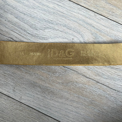 The Dolce &amp; Gabbana 2000’s Gold Tone D&amp;G Logo Belt is made from metallic gold leather and features a large D&amp;G logo buckle shaped directly as the brand’s initials. The buckle uses a cursive D&amp;G logo set over a glittery gold background, framed by a solid gold-tone metal outline. The entire buckle and belt maintain a consistent gold color scheme typical of Dolce &amp; Gabbana accessories from the 2000s.