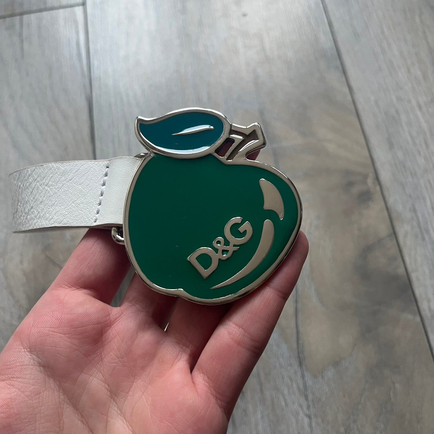 Archive Dolce &amp; Gabbana belt from the 2000s featuring a pretty big metal apple-shaped buckle with a leaf detail, finished in green with silver accents and a small D&amp;G logo. The belt is crafted from white leather with a silver-toned interior, includes a small metal D&amp;G logo at the tip, and is marked Made in Romania. Comes with original tags; the oversized buckle contrasts with the slim belt width.