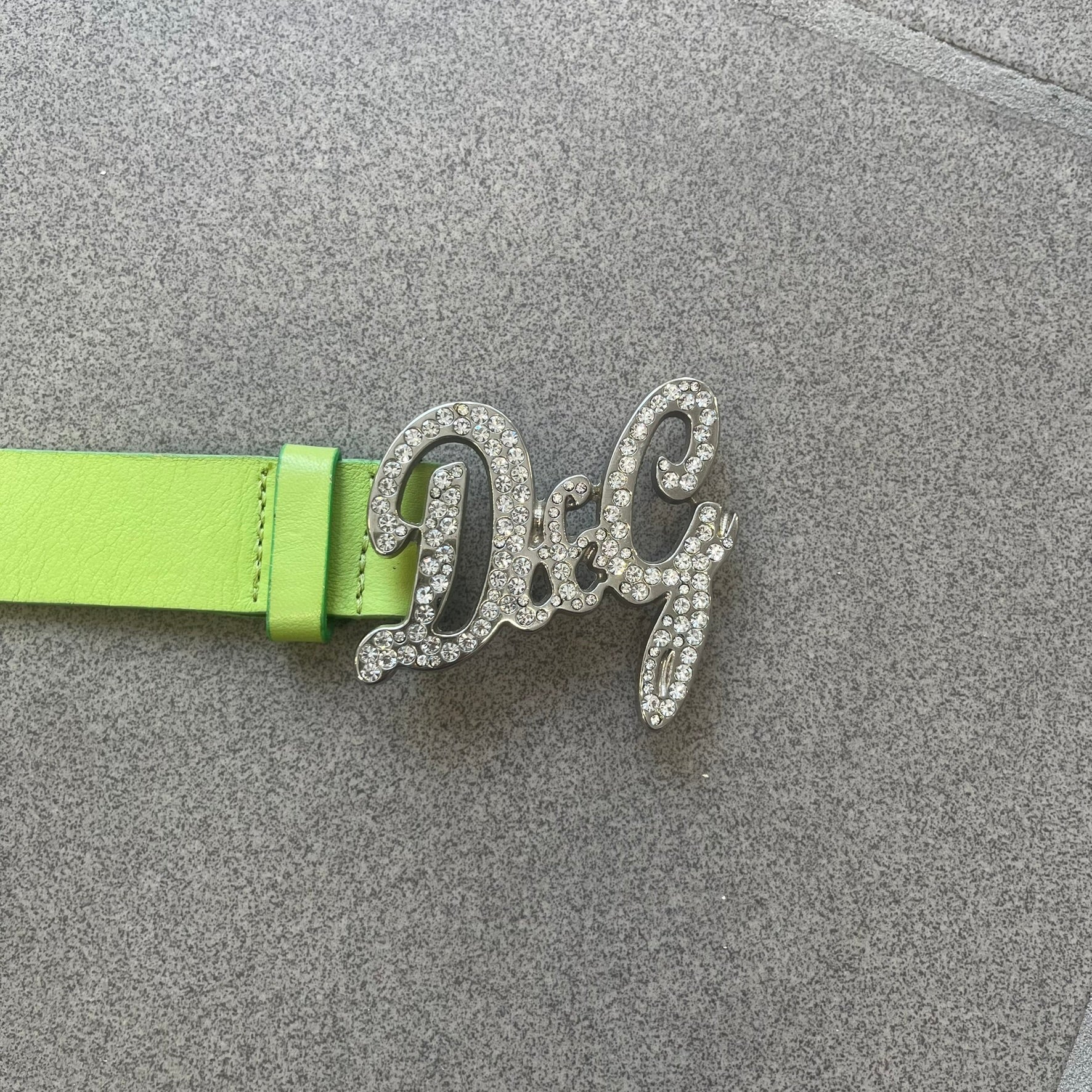 Dolce &amp; Gabbana 2000s Green Crystal Buckle Belt features a lime green leather strap with a stainless steel D&amp;G logo buckle. The buckle has a decorative italic-style font and is fully covered with crystal/rhinestone detailing on the front. Made in Italy.