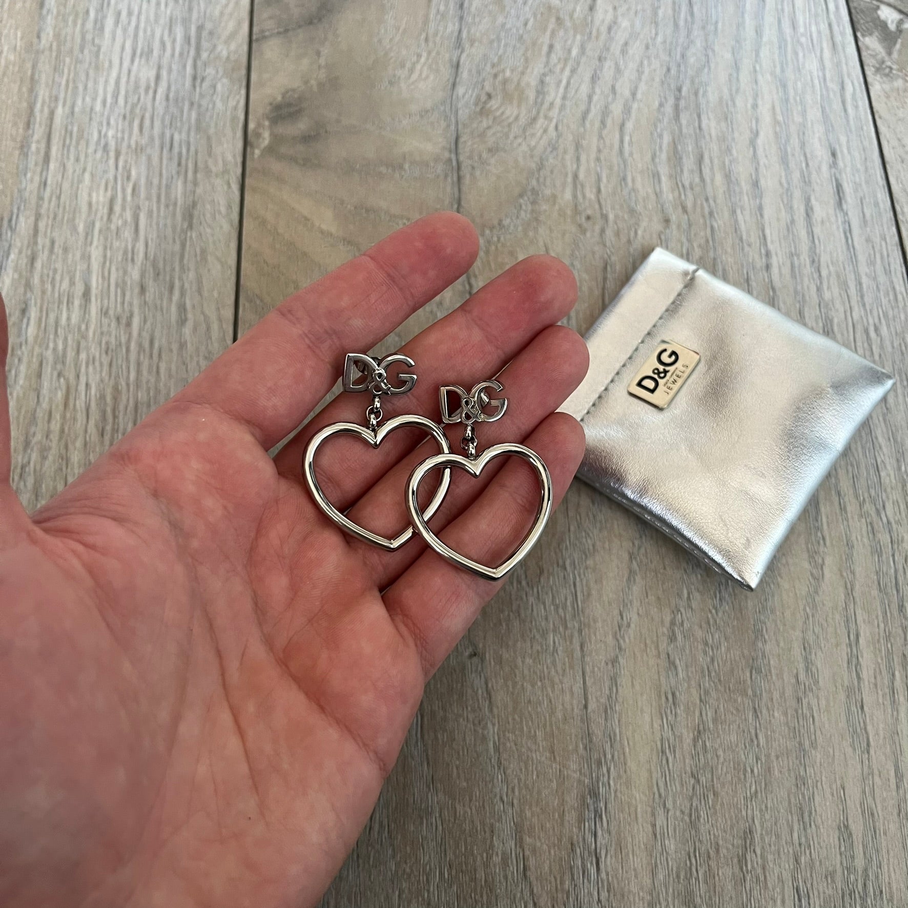 Dolce & Gabbana DJ1013 2000s Heart Earrings feature a silver-tone dangling design with D&G logo studs at the top and large outlined heart charms suspended below. The heart pendants are movable and hang freely from the logo attachment. The earrings come with the original Dolce & Gabbana dust bag.