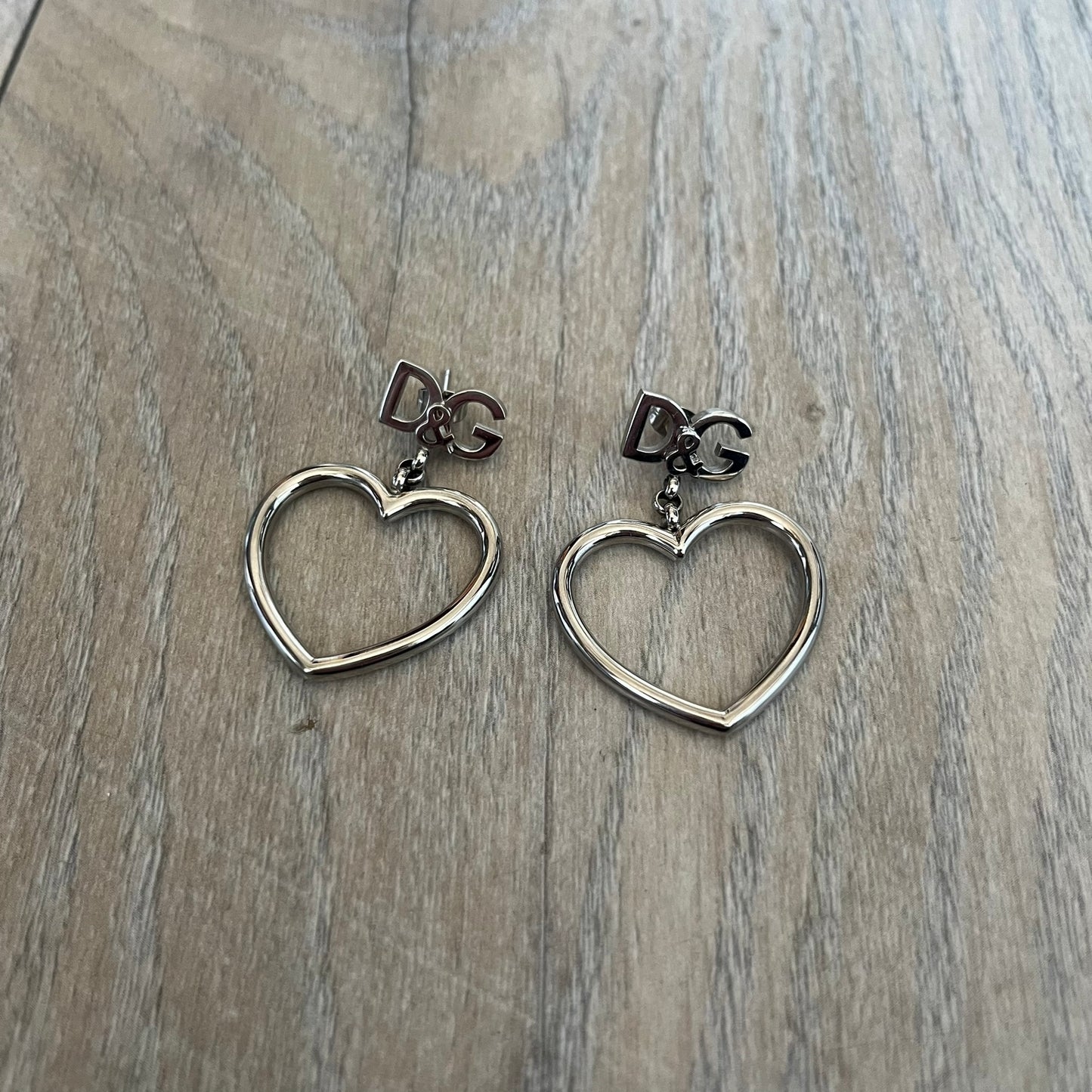 Dolce & Gabbana DJ1013 2000s Heart Earrings feature a silver-tone dangling design with D&G logo studs at the top and large outlined heart charms suspended below. The heart pendants are movable and hang freely from the logo attachment. The earrings come with the original Dolce & Gabbana dust bag.
