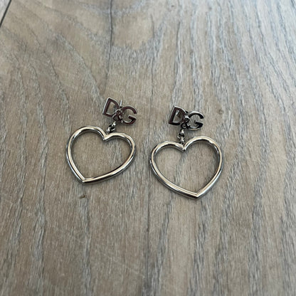 Dolce & Gabbana DJ1013 2000s Heart Earrings feature a silver-tone dangling design with D&G logo studs at the top and large outlined heart charms suspended below. The heart pendants are movable and hang freely from the logo attachment. The earrings come with the original Dolce & Gabbana dust bag.