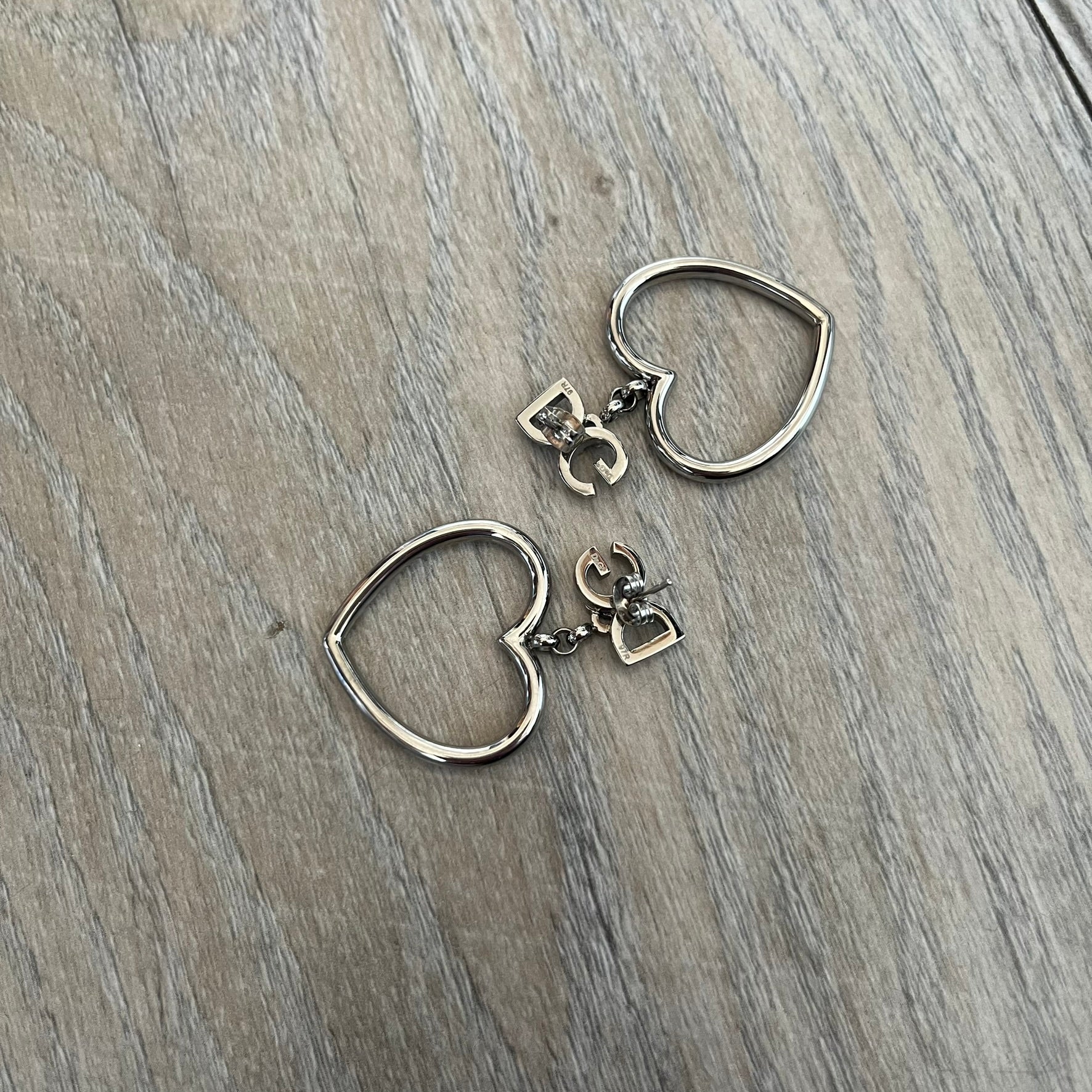 Dolce & Gabbana DJ1013 2000s Heart Earrings feature a silver-tone dangling design with D&G logo studs at the top and large outlined heart charms suspended below. The heart pendants are movable and hang freely from the logo attachment. The earrings come with the original Dolce & Gabbana dust bag.