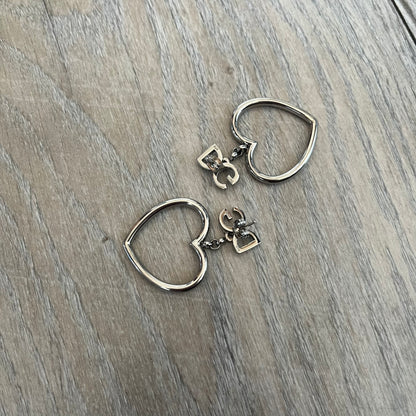 Dolce & Gabbana DJ1013 2000s Heart Earrings feature a silver-tone dangling design with D&G logo studs at the top and large outlined heart charms suspended below. The heart pendants are movable and hang freely from the logo attachment. The earrings come with the original Dolce & Gabbana dust bag.