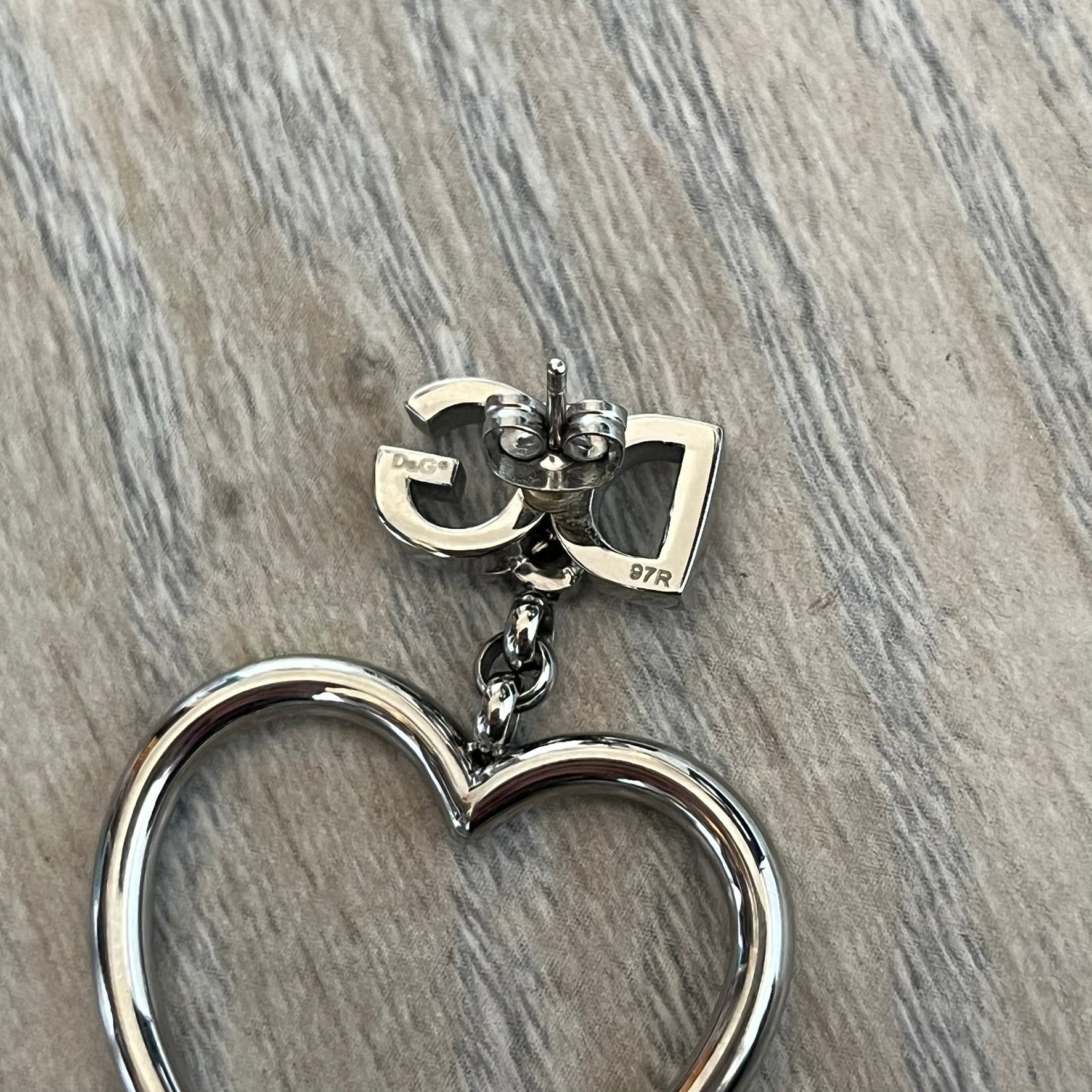 Dolce & Gabbana DJ1013 2000s Heart Earrings feature a silver-tone dangling design with D&G logo studs at the top and large outlined heart charms suspended below. The heart pendants are movable and hang freely from the logo attachment. The earrings come with the original Dolce & Gabbana dust bag.
