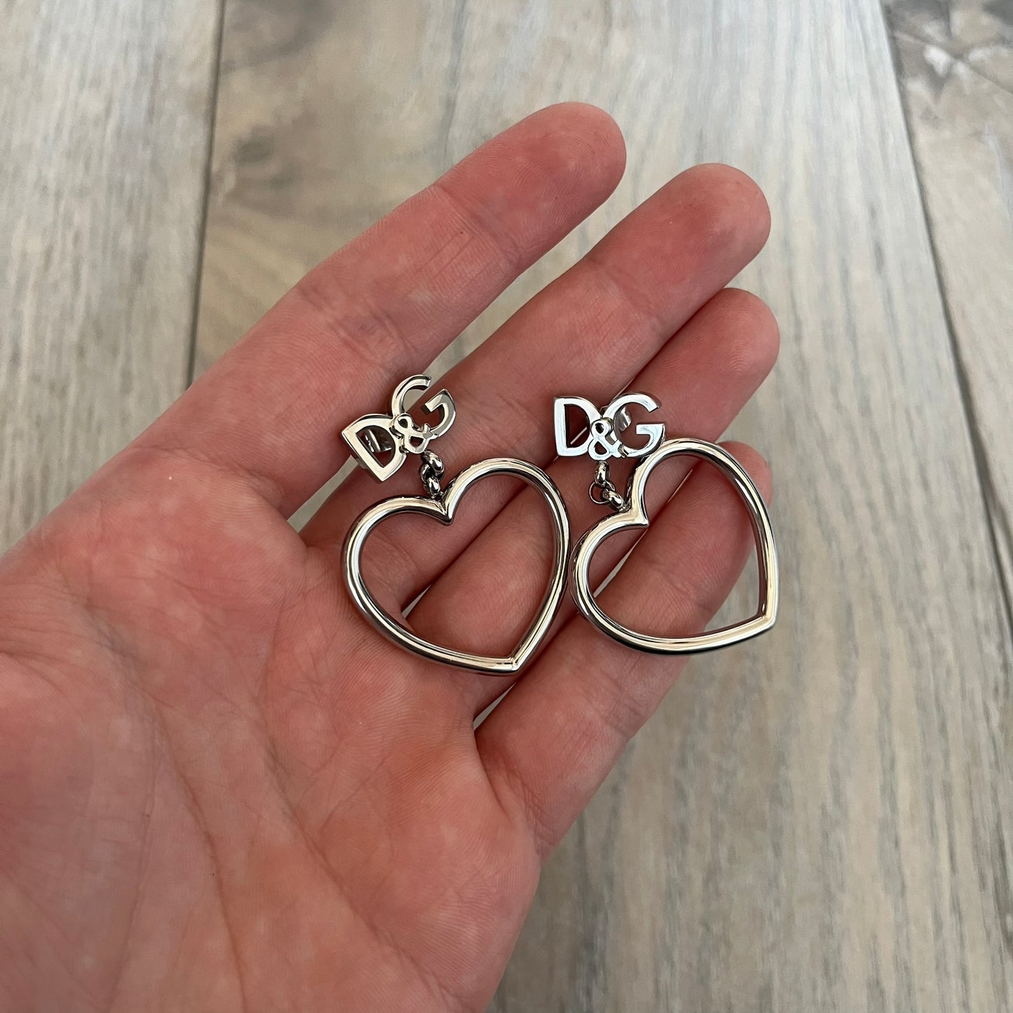 Dolce & Gabbana DJ1013 2000s Heart Earrings feature a silver-tone dangling design with D&G logo studs at the top and large outlined heart charms suspended below. The heart pendants are movable and hang freely from the logo attachment. The earrings come with the original Dolce & Gabbana dust bag.
