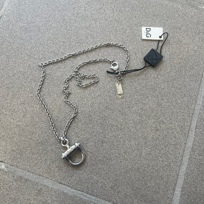 Dolce & Gabbana 2000s Horseshoe Necklace (DJ0985) is a stainless steel necklace featuring a horseshoe-shaped pendant. The pendant has a fixed section with a raised D&G logo, while the horseshoe portion is attached to it with a rotating design that allows it to spin freely. The necklace has a polished metal finish and comes from the 2000s.
