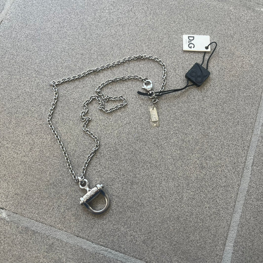 Dolce & Gabbana 2000s Horseshoe Necklace (DJ0985) is a stainless steel necklace featuring a horseshoe-shaped pendant. The pendant has a fixed section with a raised D&G logo, while the horseshoe portion is attached to it with a rotating design that allows it to spin freely. The necklace has a polished metal finish and comes from the 2000s.
