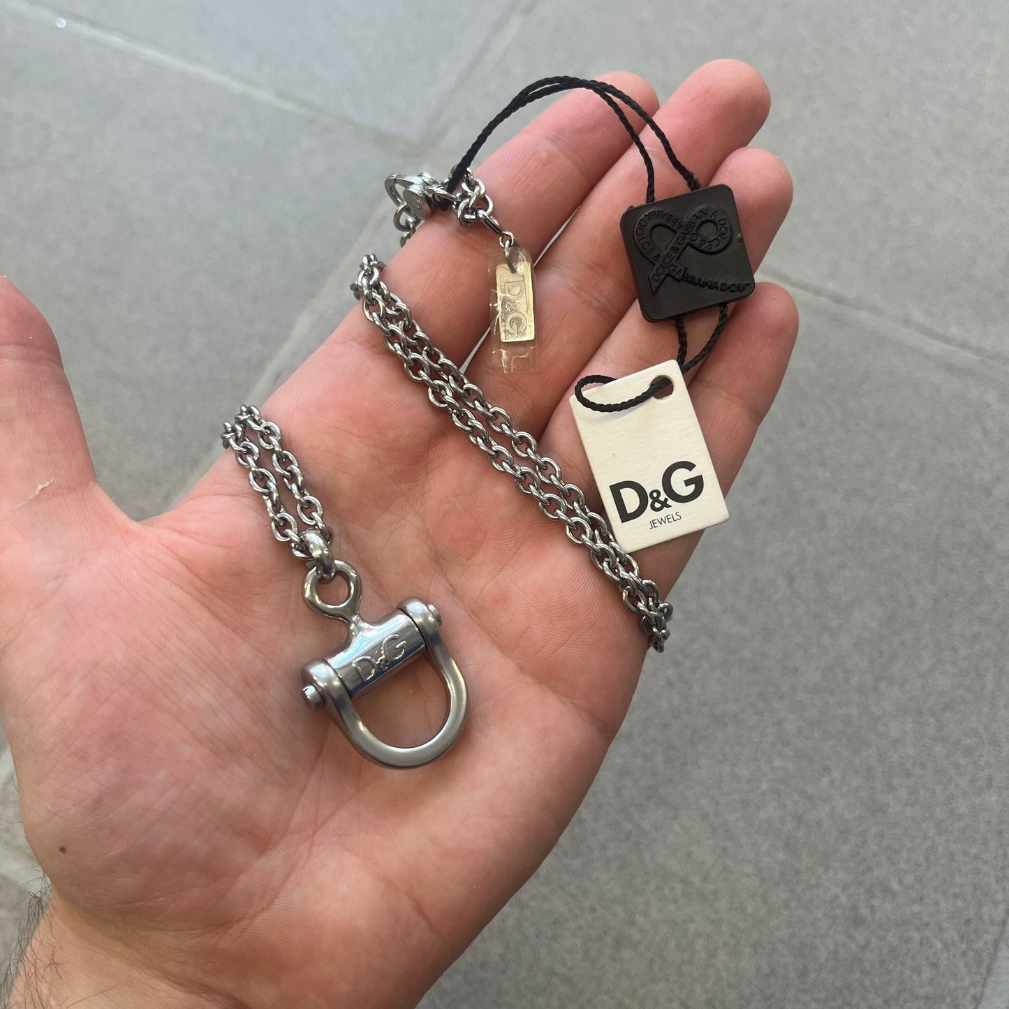 Dolce & Gabbana 2000s Horseshoe Necklace (DJ0985) is a stainless steel necklace featuring a horseshoe-shaped pendant. The pendant has a fixed section with a raised D&G logo, while the horseshoe portion is attached to it with a rotating design that allows it to spin freely. The necklace has a polished metal finish and comes from the 2000s.
