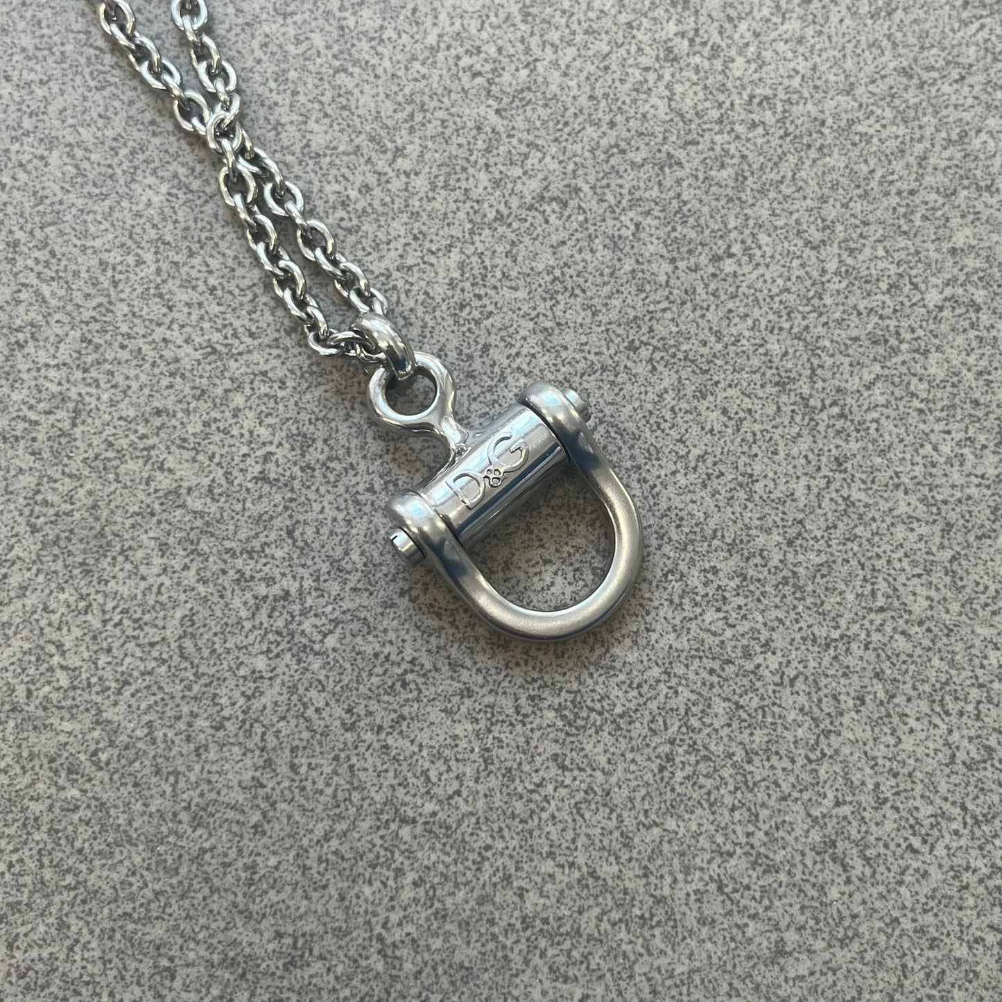 Dolce & Gabbana 2000s Horseshoe Necklace (DJ0985) is a stainless steel necklace featuring a horseshoe-shaped pendant. The pendant has a fixed section with a raised D&G logo, while the horseshoe portion is attached to it with a rotating design that allows it to spin freely. The necklace has a polished metal finish and comes from the 2000s.

