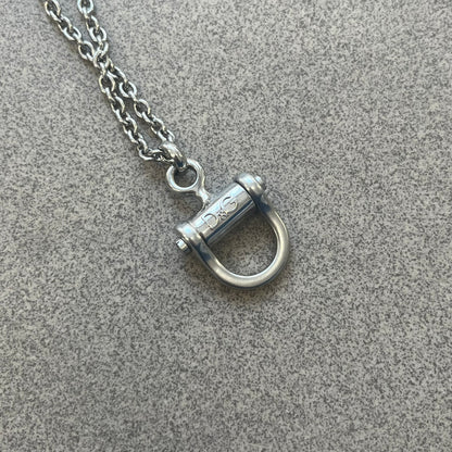 Dolce & Gabbana 2000s Horseshoe Necklace (DJ0985) is a stainless steel necklace featuring a horseshoe-shaped pendant. The pendant has a fixed section with a raised D&G logo, while the horseshoe portion is attached to it with a rotating design that allows it to spin freely. The necklace has a polished metal finish and comes from the 2000s.

