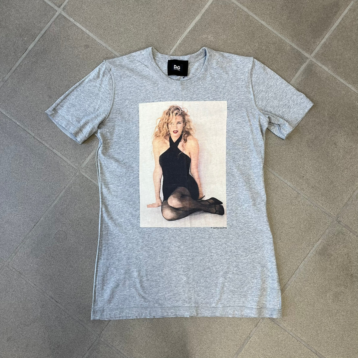 Dolce &amp; Gabbana 2000's Kim Basinger Tee is a gray T-shirt with a photo of Kim Basinger on the front and small “Herb Ritts Foundation” text below it. The lower back has D&amp;G text logo. The shirt is constructed inside-out style, with the stitching visible along the sides.