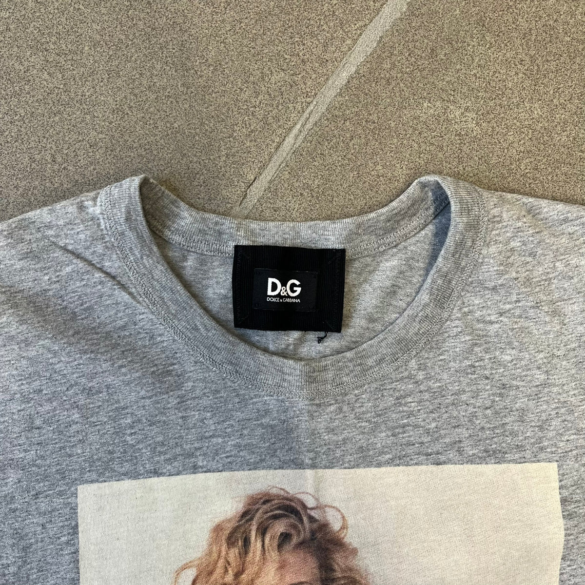 Dolce &amp; Gabbana 2000's Kim Basinger Tee is a gray T-shirt with a photo of Kim Basinger on the front and small “Herb Ritts Foundation” text below it. The lower back has D&amp;G text logo. The shirt is constructed inside-out style, with the stitching visible along the sides.