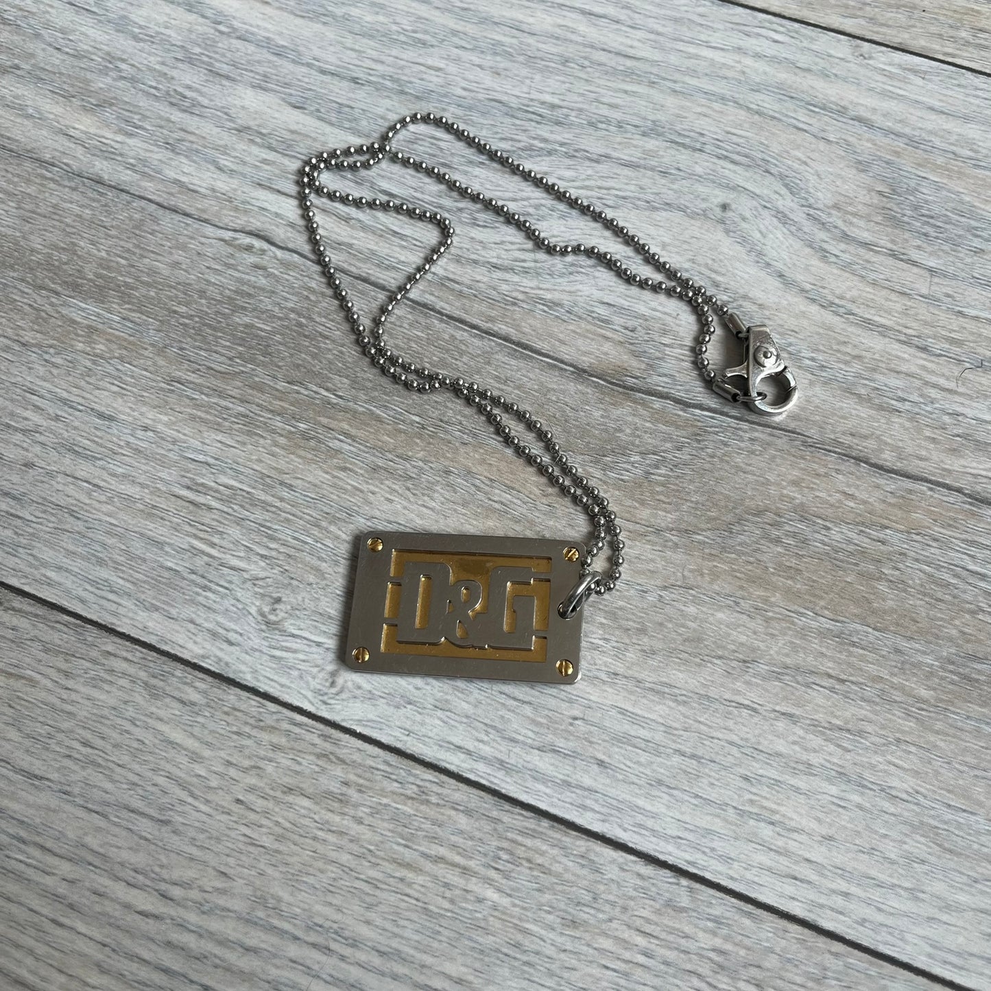 Dolce &amp; Gabbana 2000s Layered Gold/Silver Necklace DJ0721 is a chain necklace featuring a steel ball chain with lobster clasp engraved with D&amp;G branding. The pendant is constructed in a layered design with three sections: a gold-tone middle layer and silver-tone top and bottom layers. The front and back includes a cut-out D&amp;G logo with an outlined frame