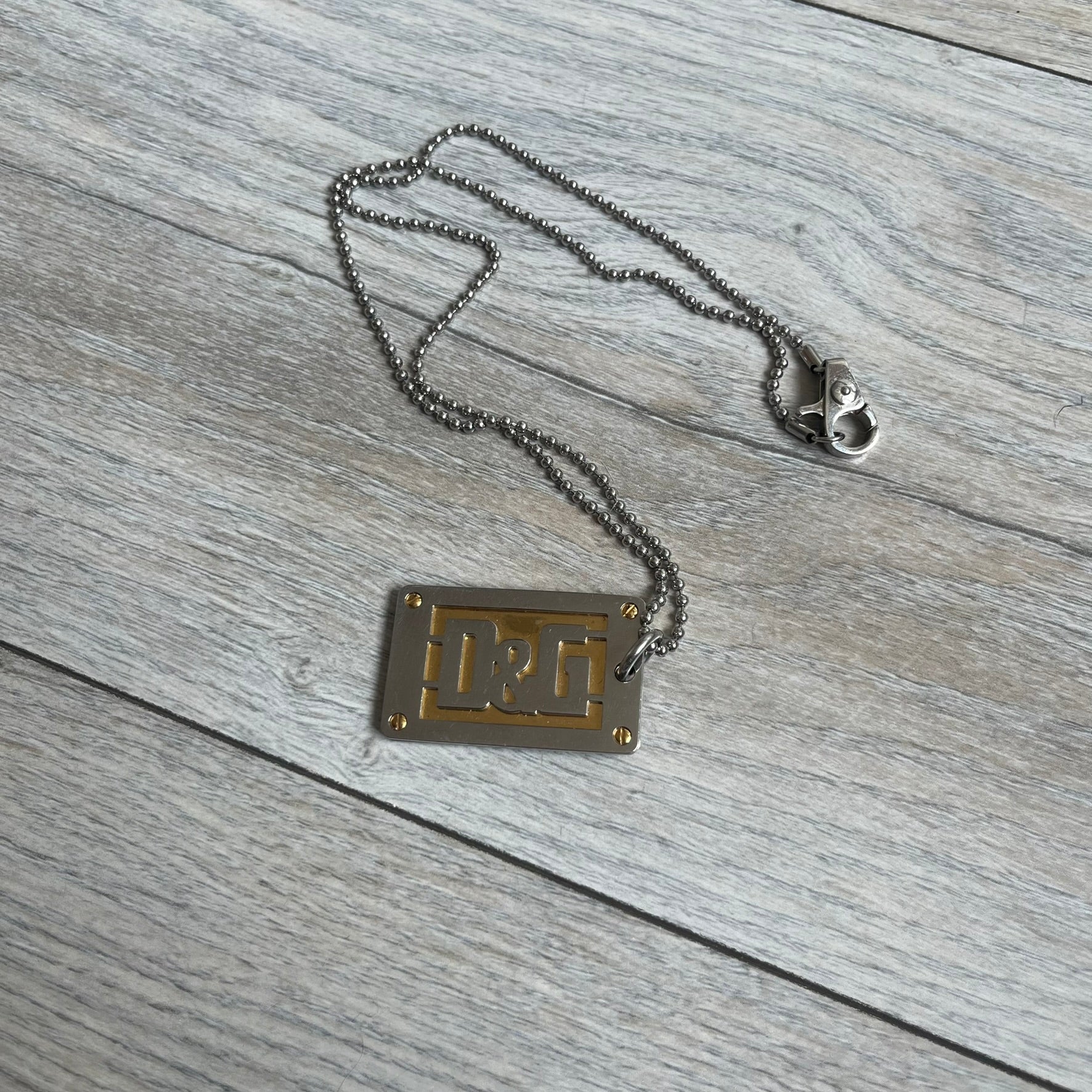 Dolce &amp; Gabbana 2000s Layered Gold/Silver Necklace DJ0721 is a chain necklace featuring a steel ball chain with lobster clasp engraved with D&amp;G branding. The pendant is constructed in a layered design with three sections: a gold-tone middle layer and silver-tone top and bottom layers. The front and back includes a cut-out D&amp;G logo with an outlined frame