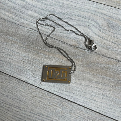 Dolce &amp; Gabbana 2000s Layered Gold/Silver Necklace DJ0721 is a chain necklace featuring a steel ball chain with lobster clasp engraved with D&amp;G branding. The pendant is constructed in a layered design with three sections: a gold-tone middle layer and silver-tone top and bottom layers. The front and back includes a cut-out D&amp;G logo with an outlined frame