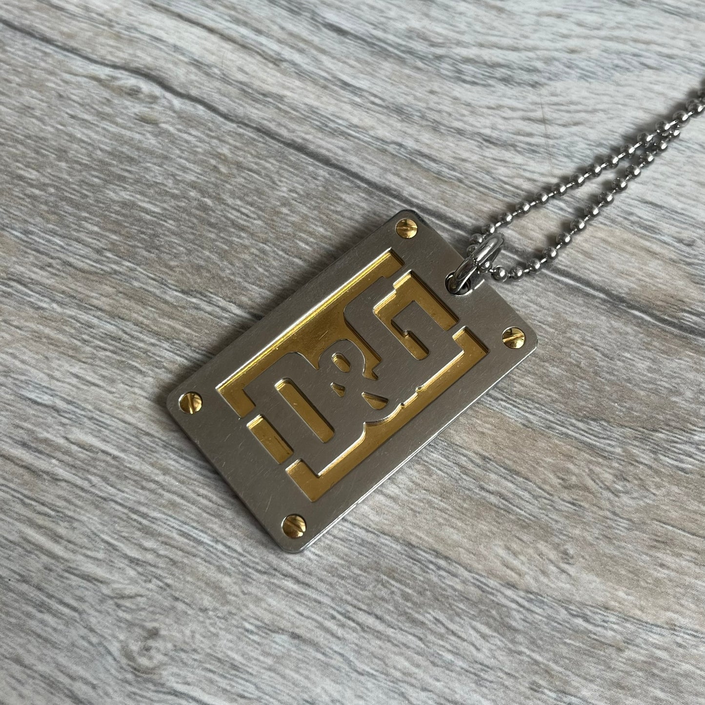 Dolce &amp; Gabbana 2000s Layered Gold/Silver Necklace DJ0721 is a chain necklace featuring a steel ball chain with lobster clasp engraved with D&amp;G branding. The pendant is constructed in a layered design with three sections: a gold-tone middle layer and silver-tone top and bottom layers. The front and back includes a cut-out D&amp;G logo with an outlined frame
