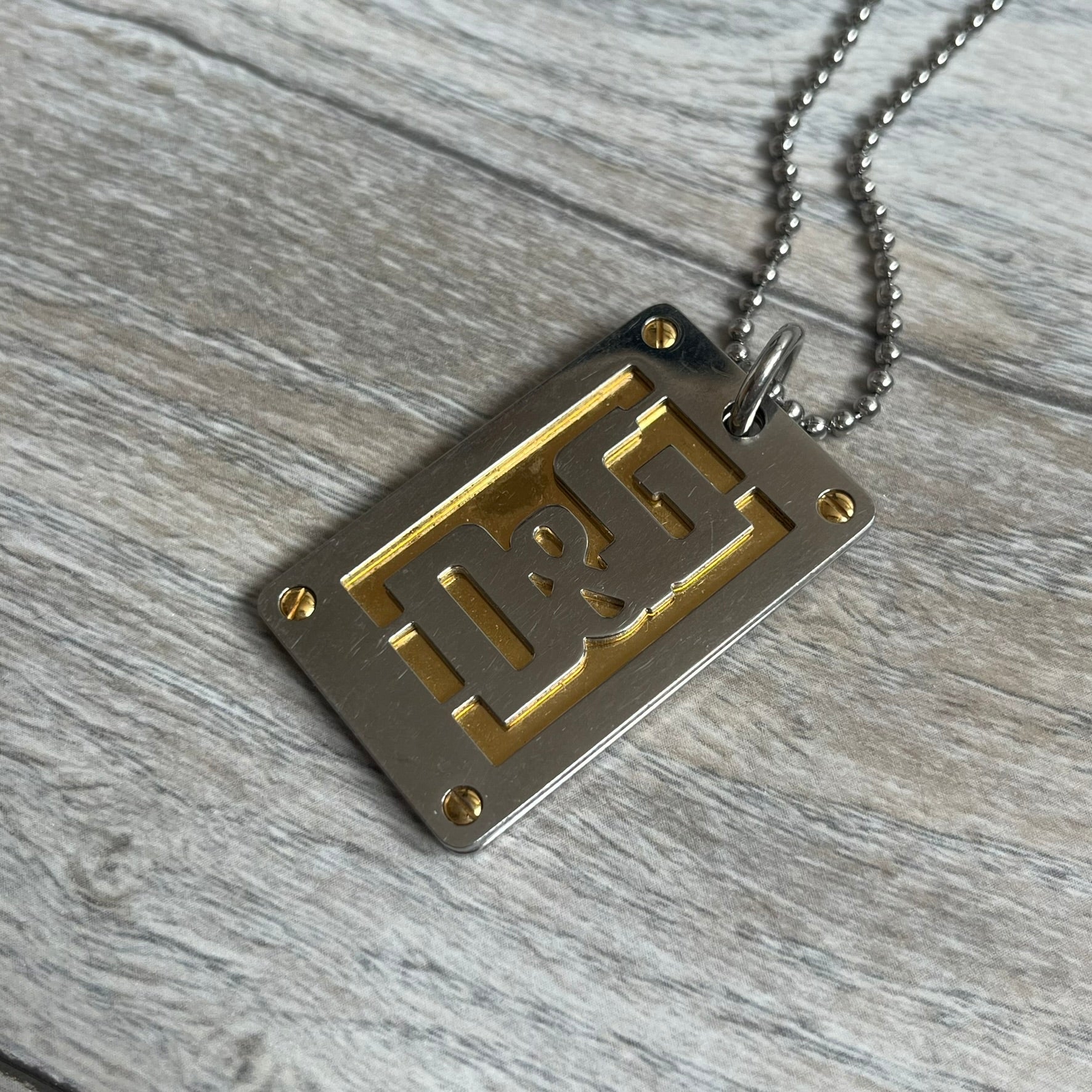 Dolce &amp; Gabbana 2000s Layered Gold/Silver Necklace DJ0721 is a chain necklace featuring a steel ball chain with lobster clasp engraved with D&amp;G branding. The pendant is constructed in a layered design with three sections: a gold-tone middle layer and silver-tone top and bottom layers. The front and back includes a cut-out D&amp;G logo with an outlined frame
