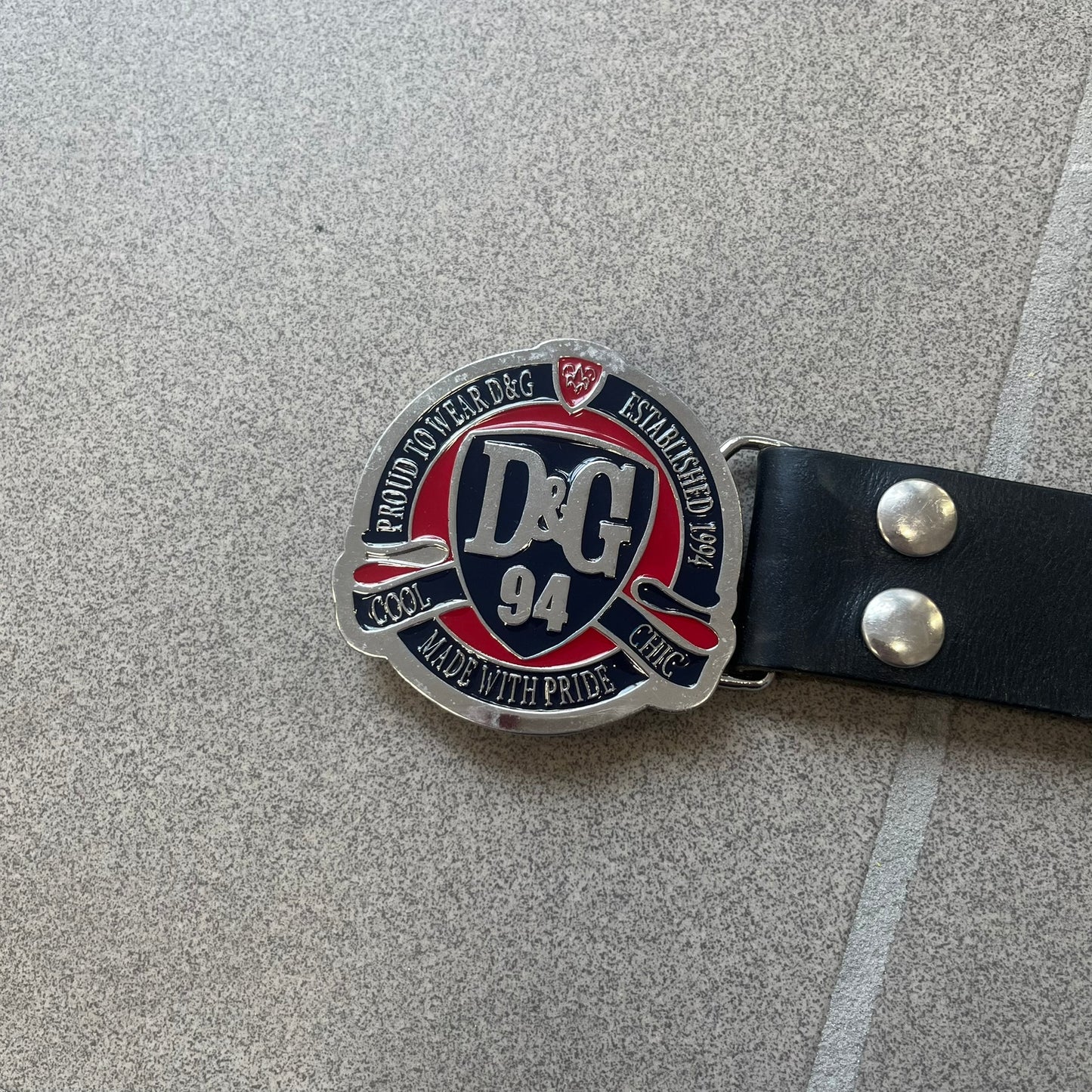 Dolce &amp; Gabbana "Made with Pride" Belt features a circular silver buckle with D&amp;G 94 at the center, surrounded by “Proud to Wear D&amp;G,” “Made with Pride,” and “Established 1994” text. Finished with subtle red and navy accents, it comes with a non-D&amp;G basic black leather belt.