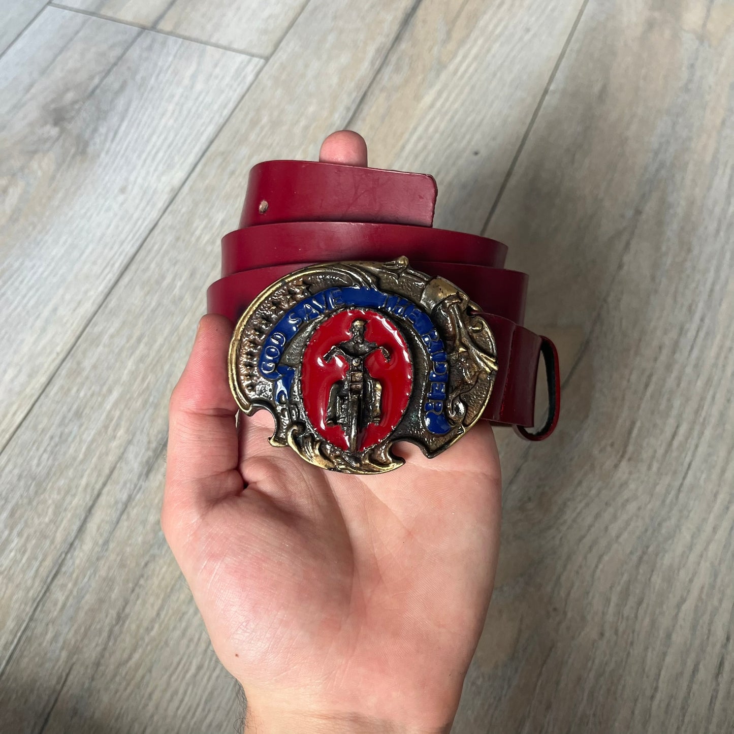 Dolce & Gabbana 2000s Moto Biker Leather Belt is a dark red leather belt made in Italy. The bronze-tone buckle features blue and red enamel detailing, with a biker graphic in the center on a red background. Above the rider is “GOD SAVE THE RIDER” text on a blue background. The buckle is finished with star detailing. Made from 100% leather.