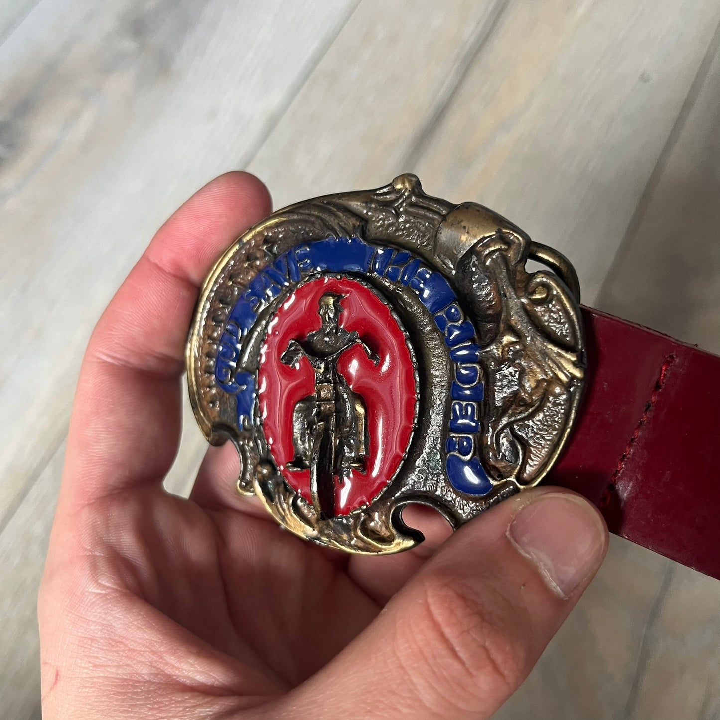 Dolce & Gabbana 2000s Moto Biker Leather Belt is a dark red leather belt made in Italy. The bronze-tone buckle features blue and red enamel detailing, with a biker graphic in the center on a red background. Above the rider is “GOD SAVE THE RIDER” text on a blue background. The buckle is finished with star detailing. Made from 100% leather.