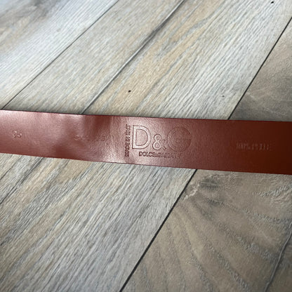 Dolce & Gabbana 2000s Moto Biker Leather Belt is a dark red leather belt made in Italy. The bronze-tone buckle features blue and red enamel detailing, with a biker graphic in the center on a red background. Above the rider is “GOD SAVE THE RIDER” text on a blue background. The buckle is finished with star detailing. Made from 100% leather.