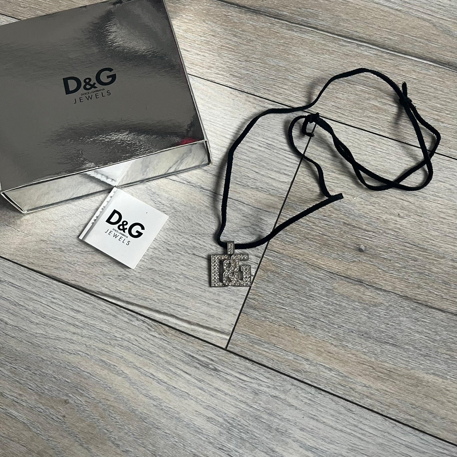 Dolce &amp; Gabbana 2000s Overlap Logo Necklace DJ0530 is a necklace featuring a pendant with overlapping D&amp;G letter cutouts in a slightly raised 3D design. The front is flooded with crystals/rhinestones. The back of the pendant is flat with a small engraved D&amp;G logo. It is attached to an adjustable cord chain with a regulator closure.
