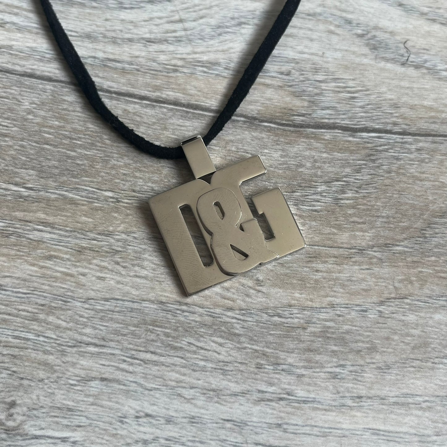 Dolce & Gabbana 2000s Overlap Logo Necklace DJ0530 is a necklace featuring a pendant with overlapping D&G letter cutouts in a slightly raised 3D design. The back of the pendant is flat with a small engraved D&G logo. It is attached to an adjustable cord chain with a regulator closure.
