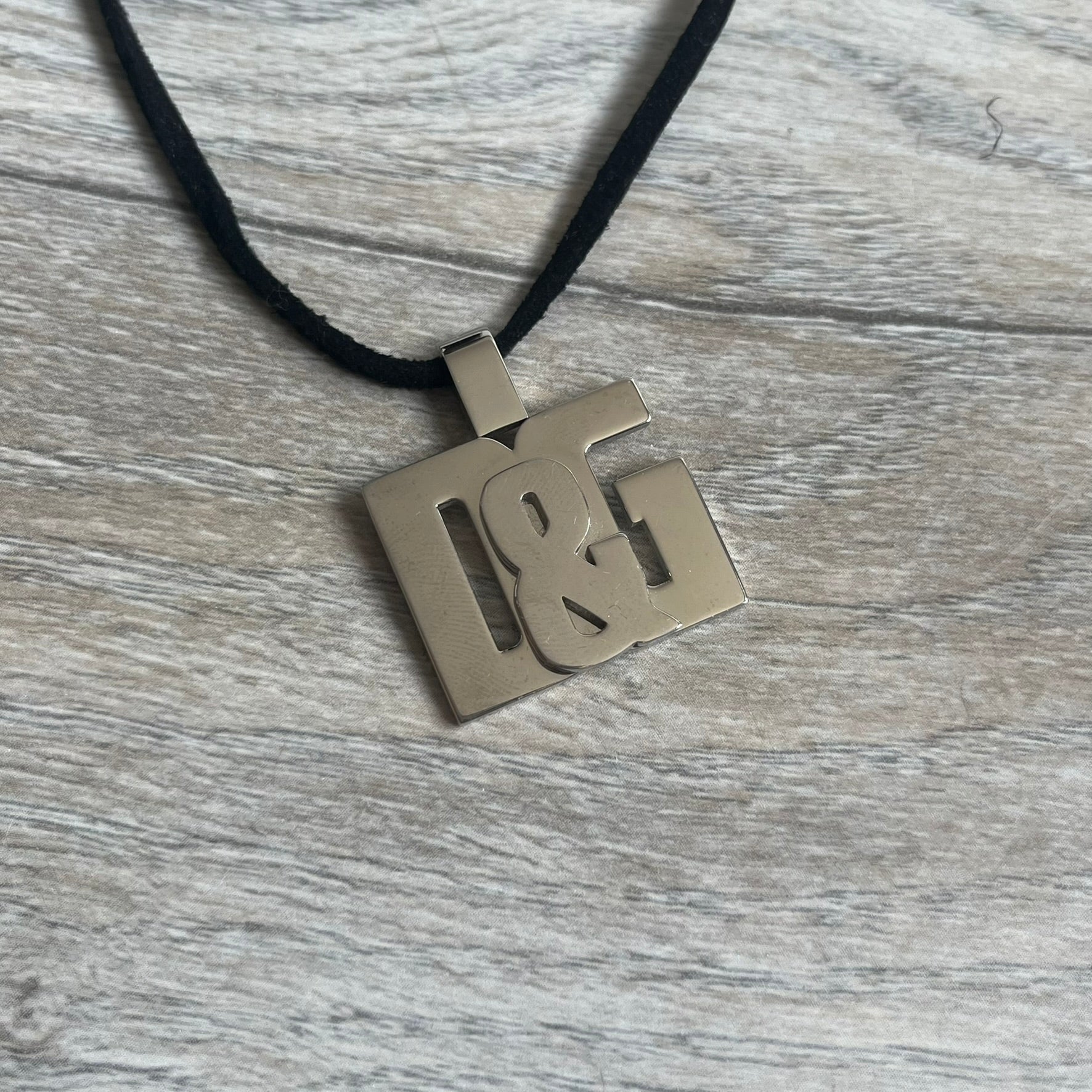 Dolce & Gabbana 2000s Overlap Logo Necklace DJ0530 is a necklace featuring a pendant with overlapping D&G letter cutouts in a slightly raised 3D design. The back of the pendant is flat with a small engraved D&G logo. It is attached to an adjustable cord chain with a regulator closure.
