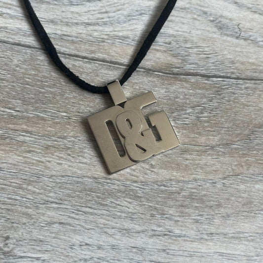 Dolce & Gabbana 2000s Overlap Logo Necklace DJ0530 is a necklace featuring a pendant with overlapping D&G letter cutouts in a slightly raised 3D design. The back of the pendant is flat with a small engraved D&G logo. It is attached to an adjustable cord chain with a regulator closure.
