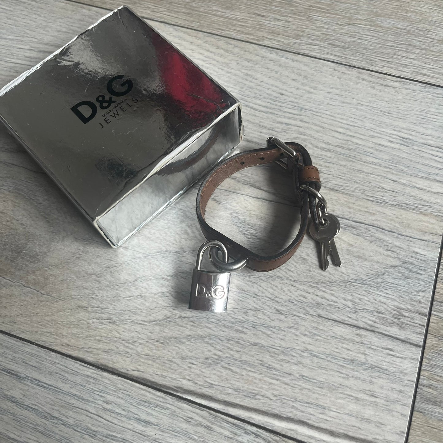 Dolce & Gabbana 2000s Padlock Leather Bracelet is a brown leather bracelet with an adjustable closure and multiple sizing holes. It features two metal loops, with a D&G engraved padlock attached to one loop and two metal keys attached to the other. The bracelet comes with its original box.
