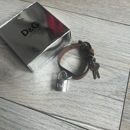 Dolce & Gabbana 2000s Padlock Leather Bracelet is a brown leather bracelet with an adjustable closure and multiple sizing holes. It features two metal loops, with a D&G engraved padlock attached to one loop and two metal keys attached to the other. The bracelet comes with its original box.
