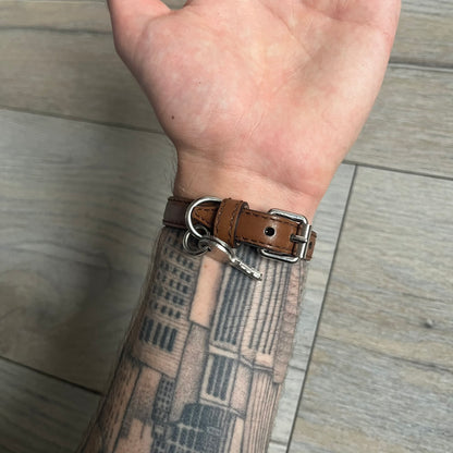 Dolce & Gabbana 2000s Padlock Leather Bracelet is a brown leather bracelet with an adjustable closure and multiple sizing holes. It features two metal loops, with a D&G engraved padlock attached to one loop and two metal keys attached to the other. The bracelet comes with its original box.
