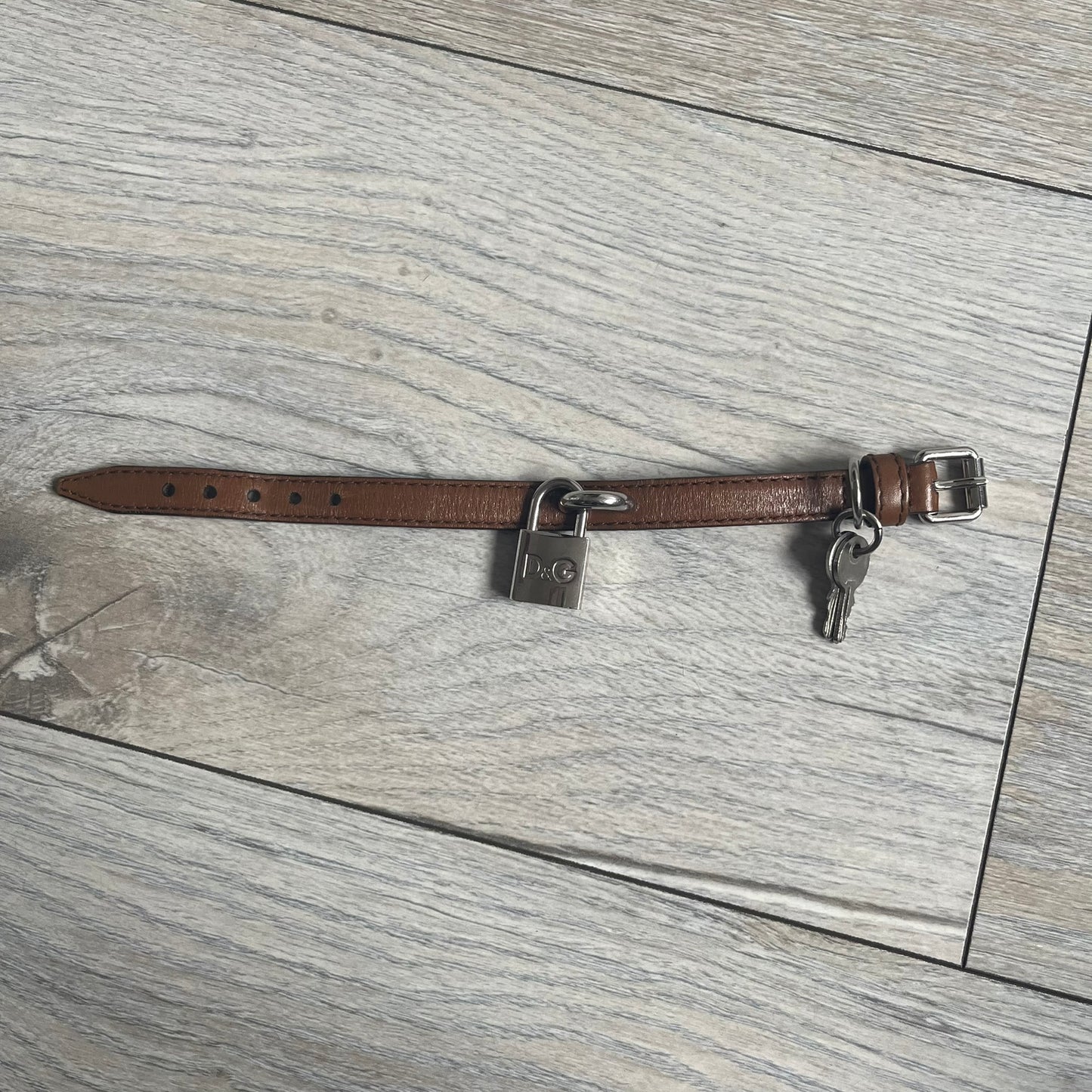 Dolce & Gabbana 2000s Padlock Leather Bracelet is a brown leather bracelet with an adjustable closure and multiple sizing holes. It features two metal loops, with a D&G engraved padlock attached to one loop and two metal keys attached to the other. The bracelet comes with its original box.
