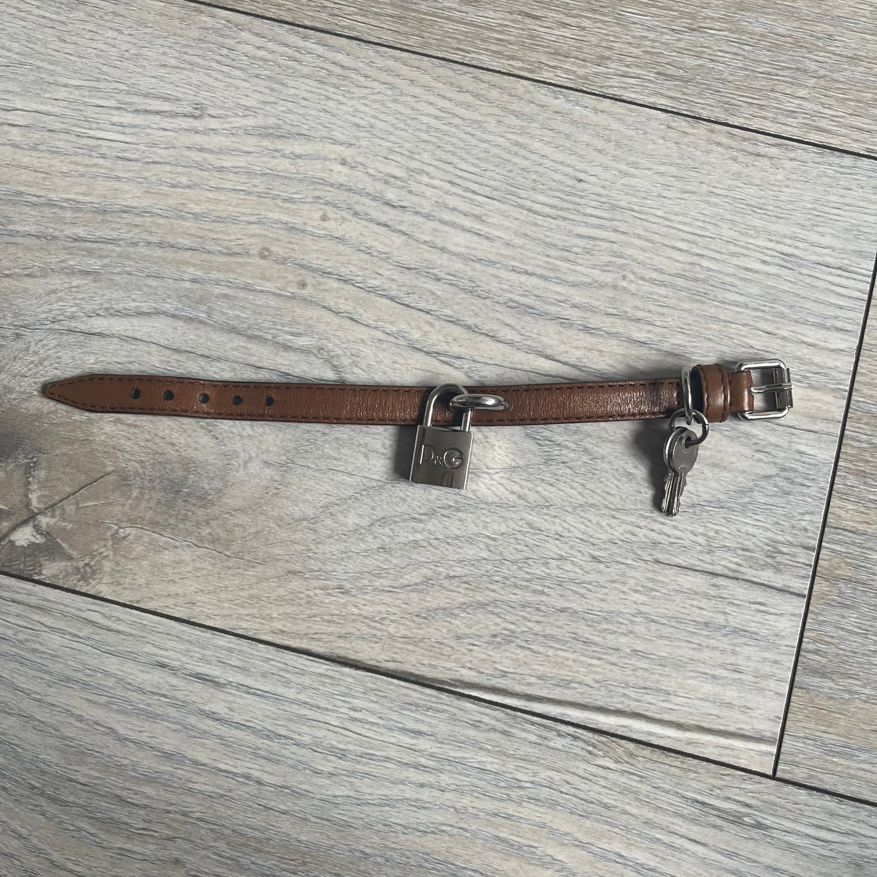 Dolce & Gabbana 2000s Padlock Leather Bracelet is a brown leather bracelet with an adjustable closure and multiple sizing holes. It features two metal loops, with a D&G engraved padlock attached to one loop and two metal keys attached to the other. The bracelet comes with its original box.
