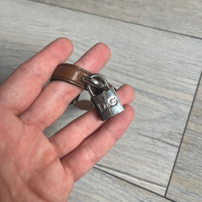 Dolce & Gabbana 2000s Padlock Leather Bracelet is a brown leather bracelet with an adjustable closure and multiple sizing holes. It features two metal loops, with a D&G engraved padlock attached to one loop and two metal keys attached to the other. The bracelet comes with its original box.
