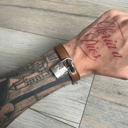 Dolce & Gabbana 2000s Padlock Leather Bracelet is a brown leather bracelet with an adjustable closure and multiple sizing holes. It features two metal loops, with a D&G engraved padlock attached to one loop and two metal keys attached to the other. The bracelet comes with its original box.
