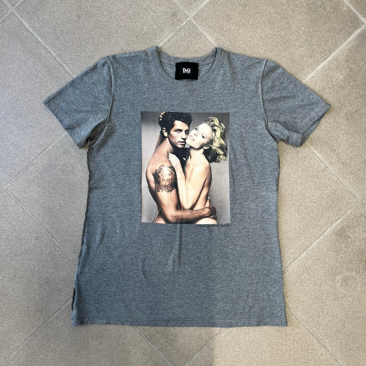 ● Dolce &amp; Gabbana 2000's Photo by Mario Testino Tee is a gray T-shirt with a photo of Naked Man and Woman holding each other on the front, and small “Photo by Mario Testino” text below it. The upper back has “D&amp;G 10 La Roue De La Fortune” text. The shirt is constructed inside-out style, with the stitching visible along the sides.