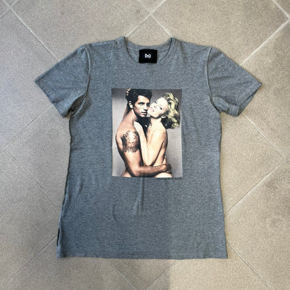 ● Dolce &amp; Gabbana 2000's Photo by Mario Testino Tee is a gray T-shirt with a photo of Naked Man and Woman holding each other on the front, and small “Photo by Mario Testino” text below it. The upper back has “D&amp;G 10 La Roue De La Fortune” text. The shirt is constructed inside-out style, with the stitching visible along the sides.