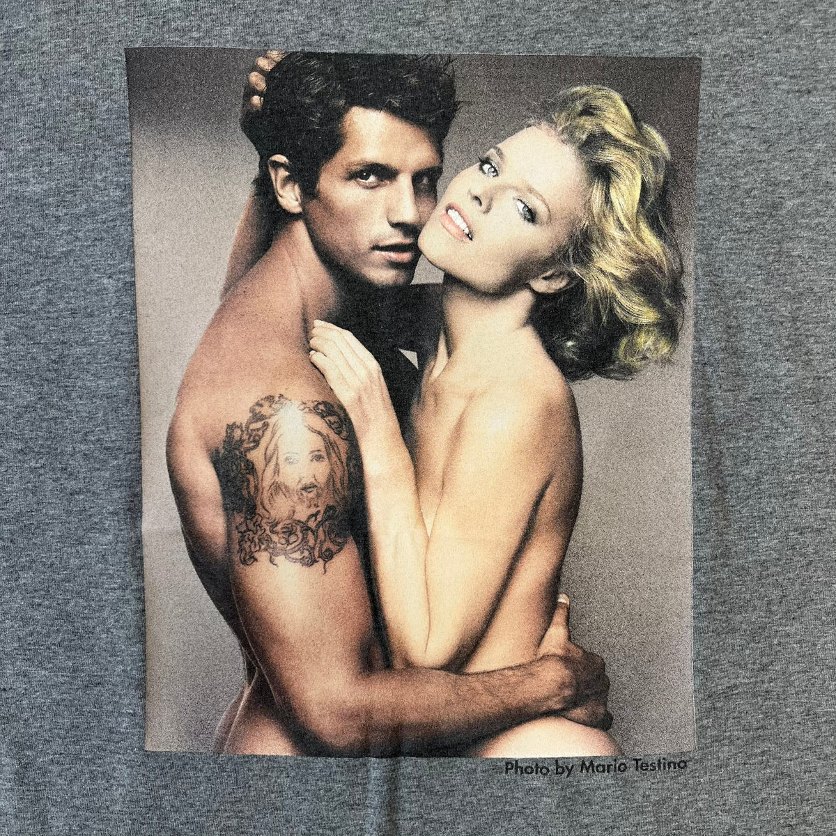 ● Dolce &amp; Gabbana 2000's Photo by Mario Testino Tee is a gray T-shirt with a photo of Naked Man and Woman holding each other on the front, and small “Photo by Mario Testino” text below it. The upper back has “D&amp;G 10 La Roue De La Fortune” text. The shirt is constructed inside-out style, with the stitching visible along the sides.