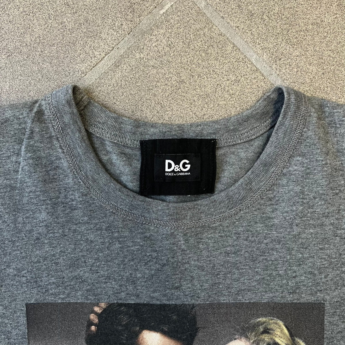 ● Dolce &amp; Gabbana 2000's Photo by Mario Testino Tee is a gray T-shirt with a photo of Naked Man and Woman holding each other on the front, and small “Photo by Mario Testino” text below it. The upper back has “D&amp;G 10 La Roue De La Fortune” text. The shirt is constructed inside-out style, with the stitching visible along the sides.