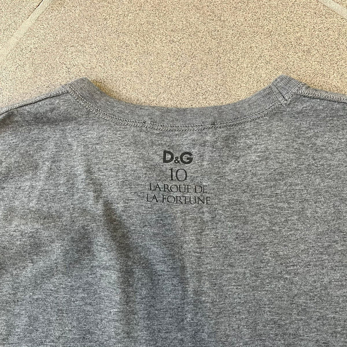 Dolce &amp; Gabbana 2000's Photo by Mario Testino Tee is a gray T-shirt with a photo of Naked Man and Woman holding each other on the front, and small “Photo by Mario Testino” text below it. The upper back has “D&amp;G 10 La Roue De La Fortune” text. The shirt is constructed inside-out style, with the stitching visible along the sides.