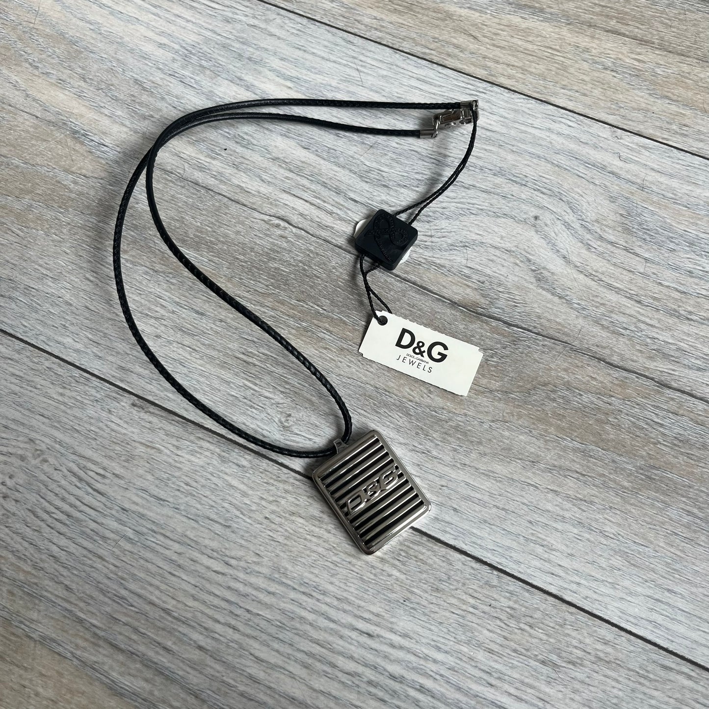Dolce & Gabbana 2000s Radiator Necklace DJ0705 is a necklace featuring a square-link chain combined with a black leather cord. The pendant is made from stainless steel with a square frame design that includes black and silver line detailing resembling a radiator structure. A raised D&G logo is positioned in the center of the ID plate, while the back is black, visible through the open metal line design.