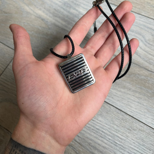 Dolce & Gabbana 2000s Radiator Necklace DJ0705 is a necklace featuring a square-link chain combined with a black leather cord. The pendant is made from stainless steel with a square frame design that includes black and silver line detailing resembling a radiator structure. A raised D&G logo is positioned in the center of the ID plate, while the back is black, visible through the open metal line design.