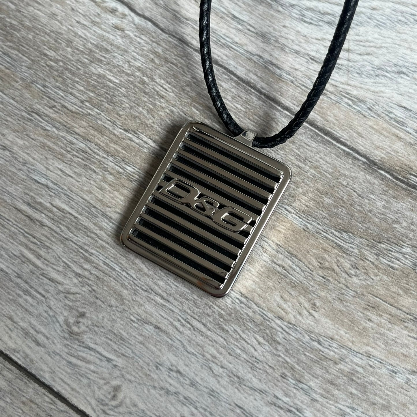 Dolce & Gabbana 2000s Radiator Necklace DJ0705 is a necklace featuring a square-link chain combined with a black leather cord. The pendant is made from stainless steel with a square frame design that includes black and silver line detailing resembling a radiator structure. A raised D&G logo is positioned in the center of the ID plate, while the back is black, visible through the open metal line design.
