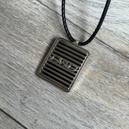Dolce & Gabbana 2000s Radiator Necklace DJ0705 is a necklace featuring a square-link chain combined with a black leather cord. The pendant is made from stainless steel with a square frame design that includes black and silver line detailing resembling a radiator structure. A raised D&G logo is positioned in the center of the ID plate, while the back is black, visible through the open metal line design.