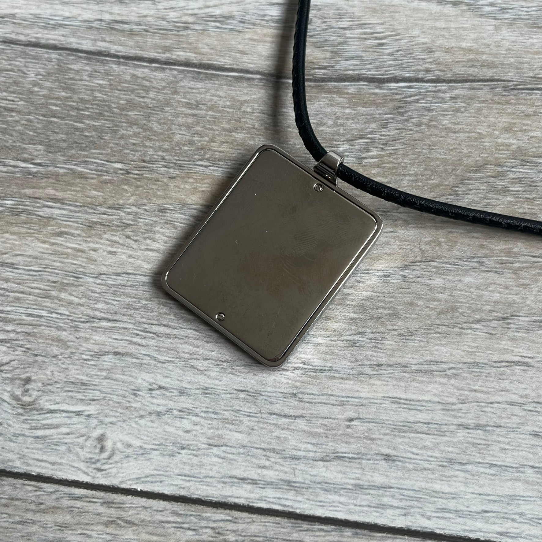 Dolce & Gabbana 2000s Radiator Necklace DJ0705 is a necklace featuring a square-link chain combined with a black leather cord. The pendant is made from stainless steel with a square frame design that includes black and silver line detailing resembling a radiator structure. A raised D&G logo is positioned in the center of the ID plate, while the back is black, visible through the open metal line design.