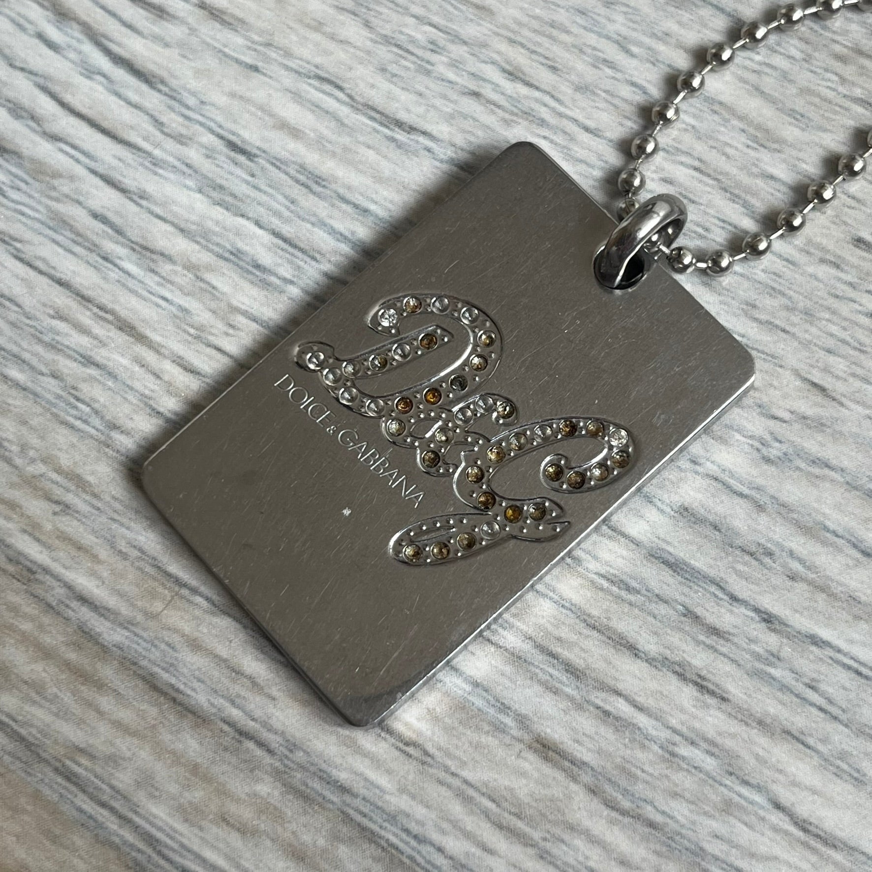 Dolce &amp; Gabbana Rhinestones Dog Tag Necklace DJ0704 crafted from polished stainless steel. Features a rectangular pendant with the iconic D&amp;G logo detailed in rhinestones, and “Dolce &amp; Gabbana” engraved subtly below. Comes with a basic non-D&amp;G steel ball chain.